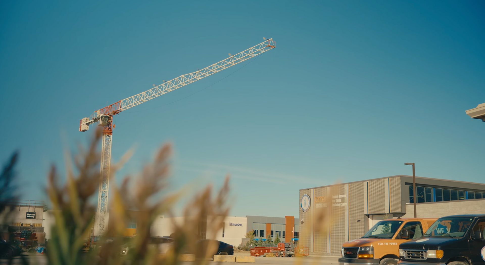 Construction crane against a blue sky, with a building and parked vehicles in the background.