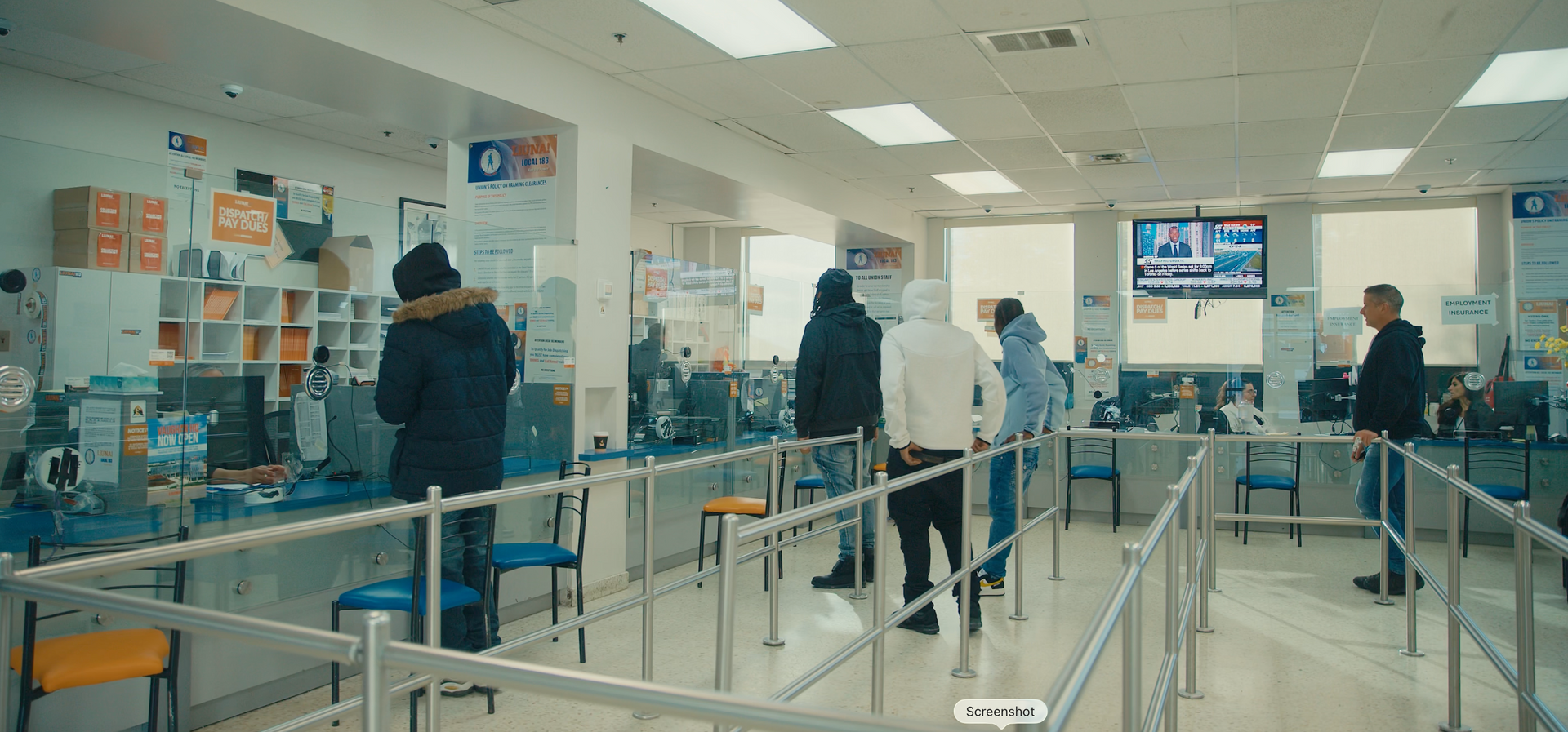 People in a line at a service counter. Interior shot, overhead lighting. Many wearing hoods and jackets. Barriers separate the line.