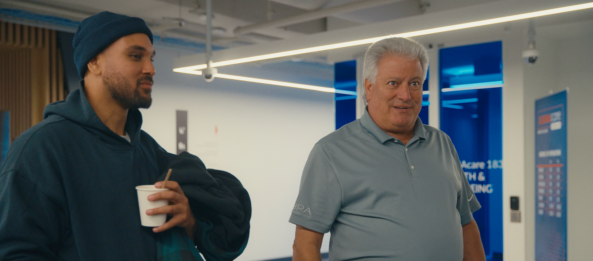 Two men inside. One in a blue beanie holds a cup. The other wears a gray shirt. They stand near a blue sign.
