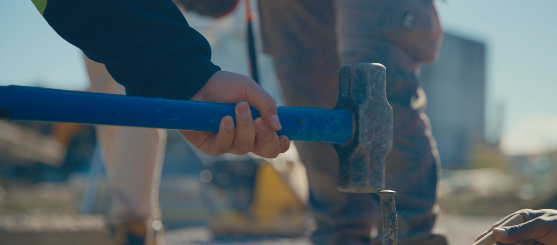 Person using a sledgehammer, focused on the hammer hitting a metal object. Blue handle, outdoor setting.