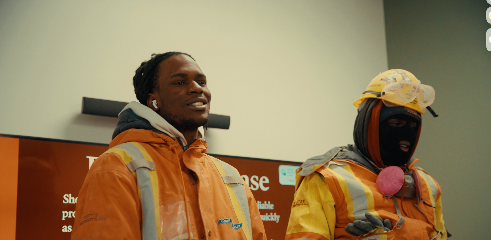 Two people in high-visibility safety gear stand in an interior space; one wears a hard hat and mask, the other is smiling.