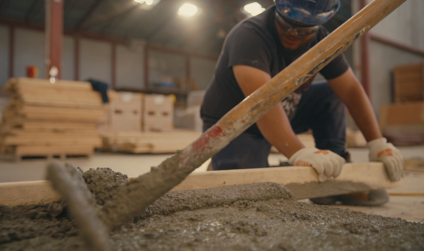 Person in safety gear using a rake to spread wet concrete on a wooden plank in a warehouse.