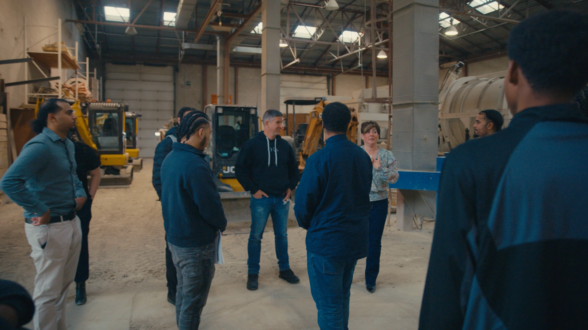 Group of people in a warehouse. They are looking at machinery, one man is talking, two small yellow tractors.
