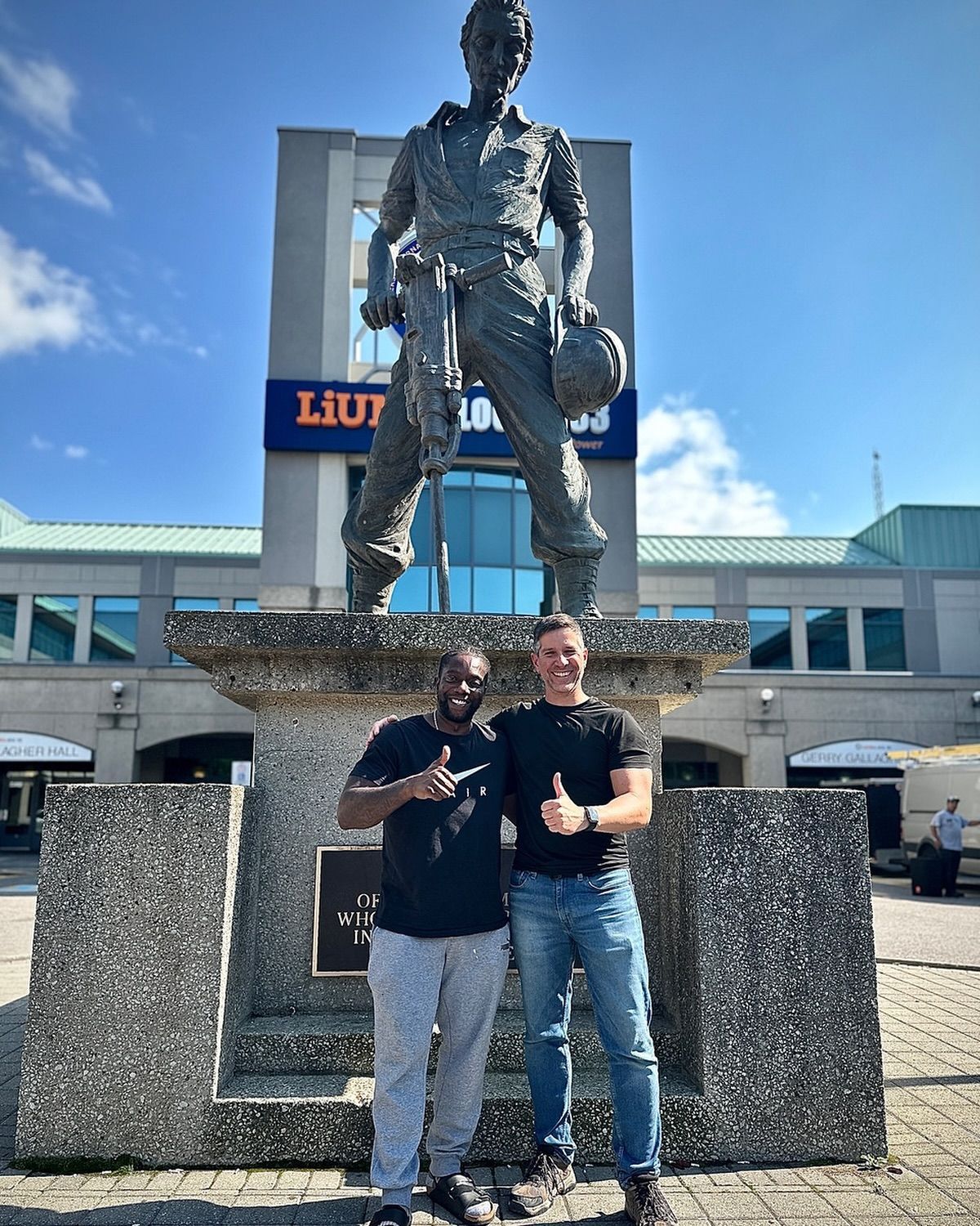 Two men are standing in front of a statue of a man holding a helmet.