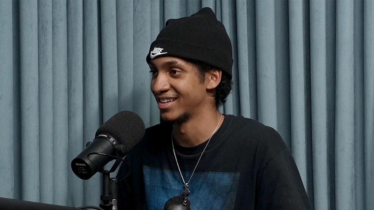 A man wearing a beanie and a necklace is standing in front of a microphone.