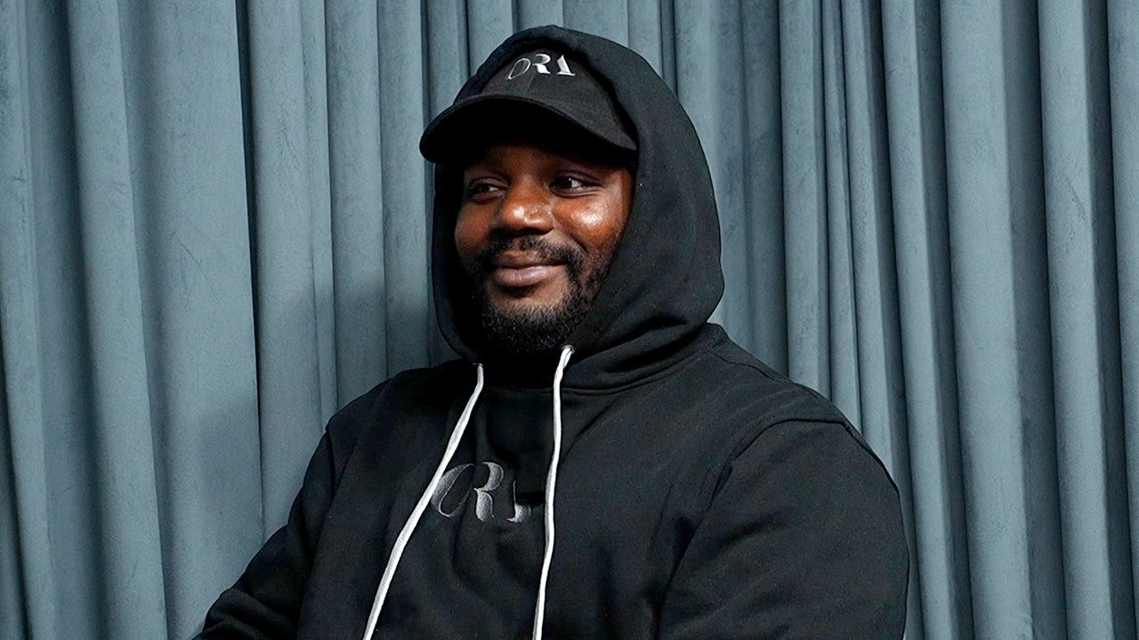 A man wearing a black hoodie and hat is sitting in front of a blue curtain.