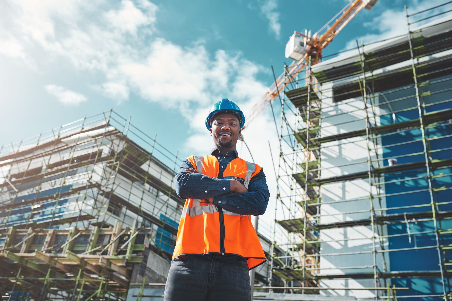 Construction worker in hard hat and vest, arms crossed, smiles in front of building under construction.