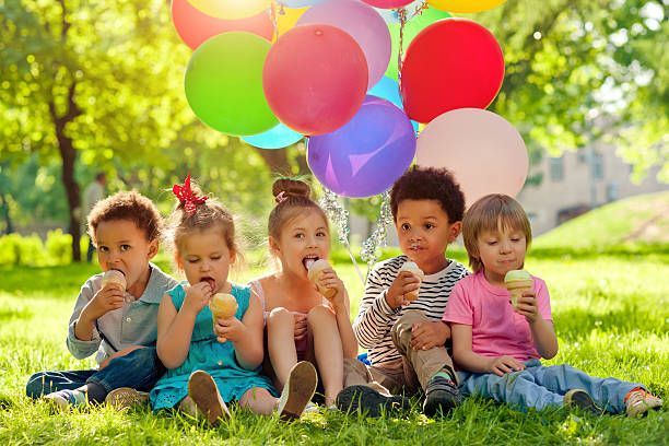 A group of children are sitting on the grass eating ice cream and holding balloons.