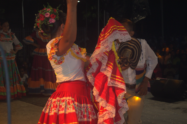Una mujer con falda roja baila con un hombre con camisa blanca. Noche, flores y trajes tradicionales.