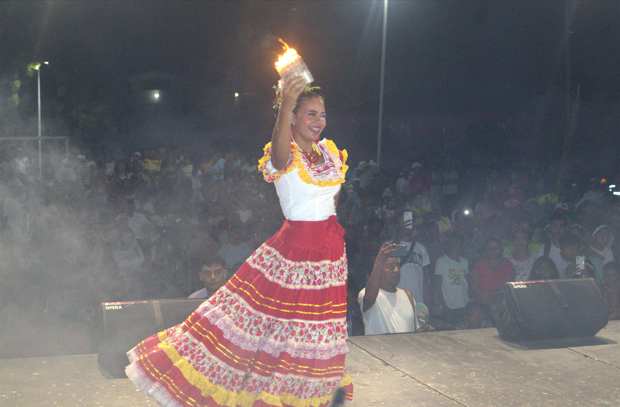 Mujer bailando en el escenario, vistiendo un vestido tradicional blanco y rojo, sosteniendo una vela encendida en la cabeza.