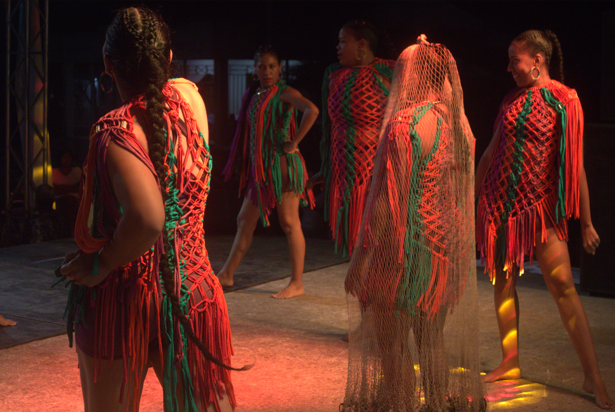 Bailarines con trajes rojos y verdes con flecos en un escenario. Uno lleva velo; otros posan, iluminados por un foco.