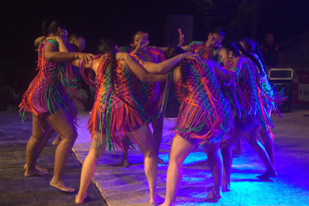 Bailarines con coloridos trajes con flecos en un círculo, iluminados por luces azules, actuando al aire libre por la noche.