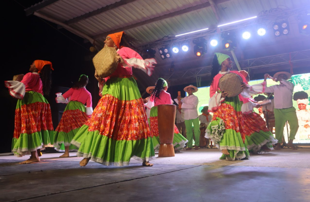 Grupo bailando en un escenario, trajes de colores brillantes: faldas verdes, blusas rosas y tambores.