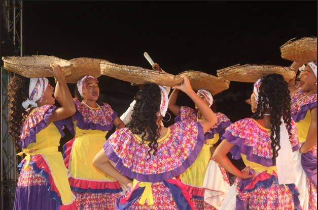 Mujeres con vestidos coloridos bailan, balanceando cestas sobre sus cabezas. Al aire libre, de noche.