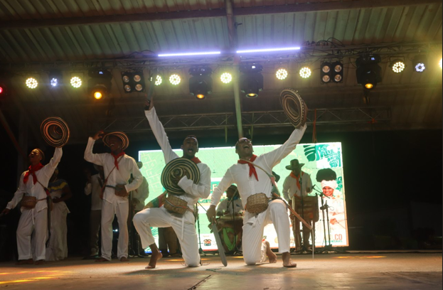 Hombres vestidos de blanco bailan y tocan instrumentos en un escenario con una pantalla al fondo.