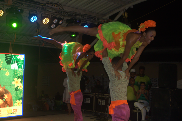 Bailarines con trajes de neón realizan elevaciones acrobáticas en el escenario. Escenario nocturno, expresiones alegres.