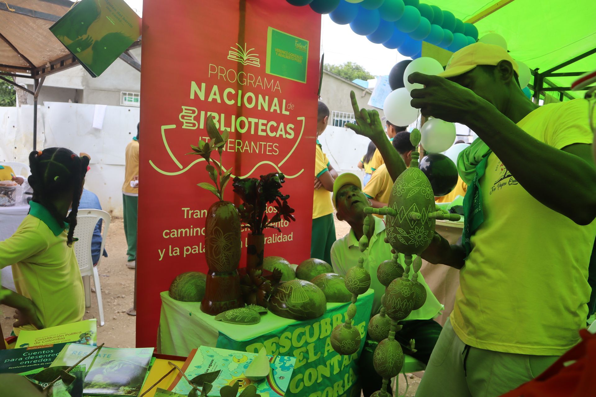 Personas en un evento al aire libre; hombre arreglando decoraciones, otros con camisetas amarillas, pancarta roja con texto, tema de biblioteca.