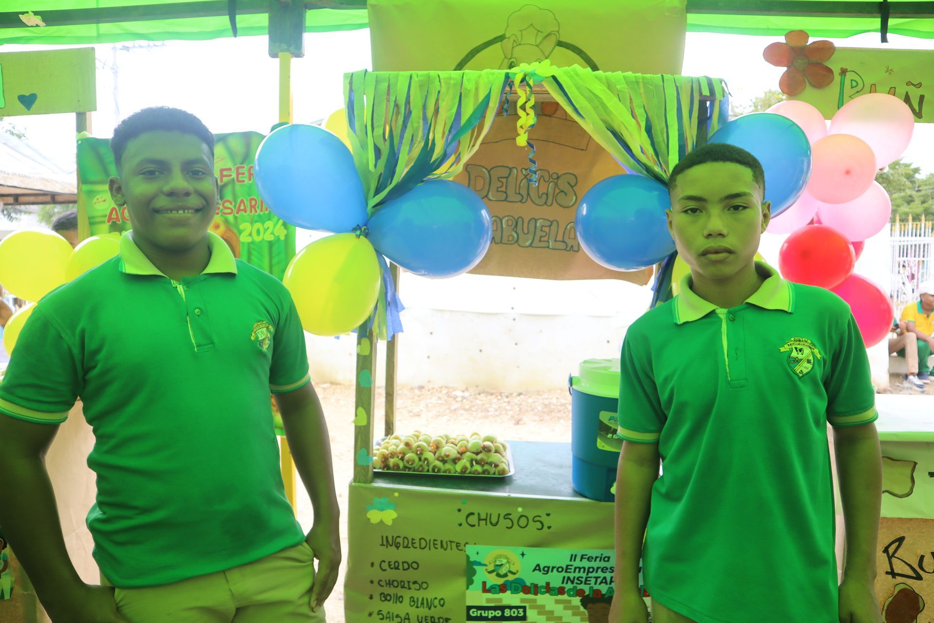 Dos niños con camisetas verdes se encuentran en un puesto decorado con comida y globos.