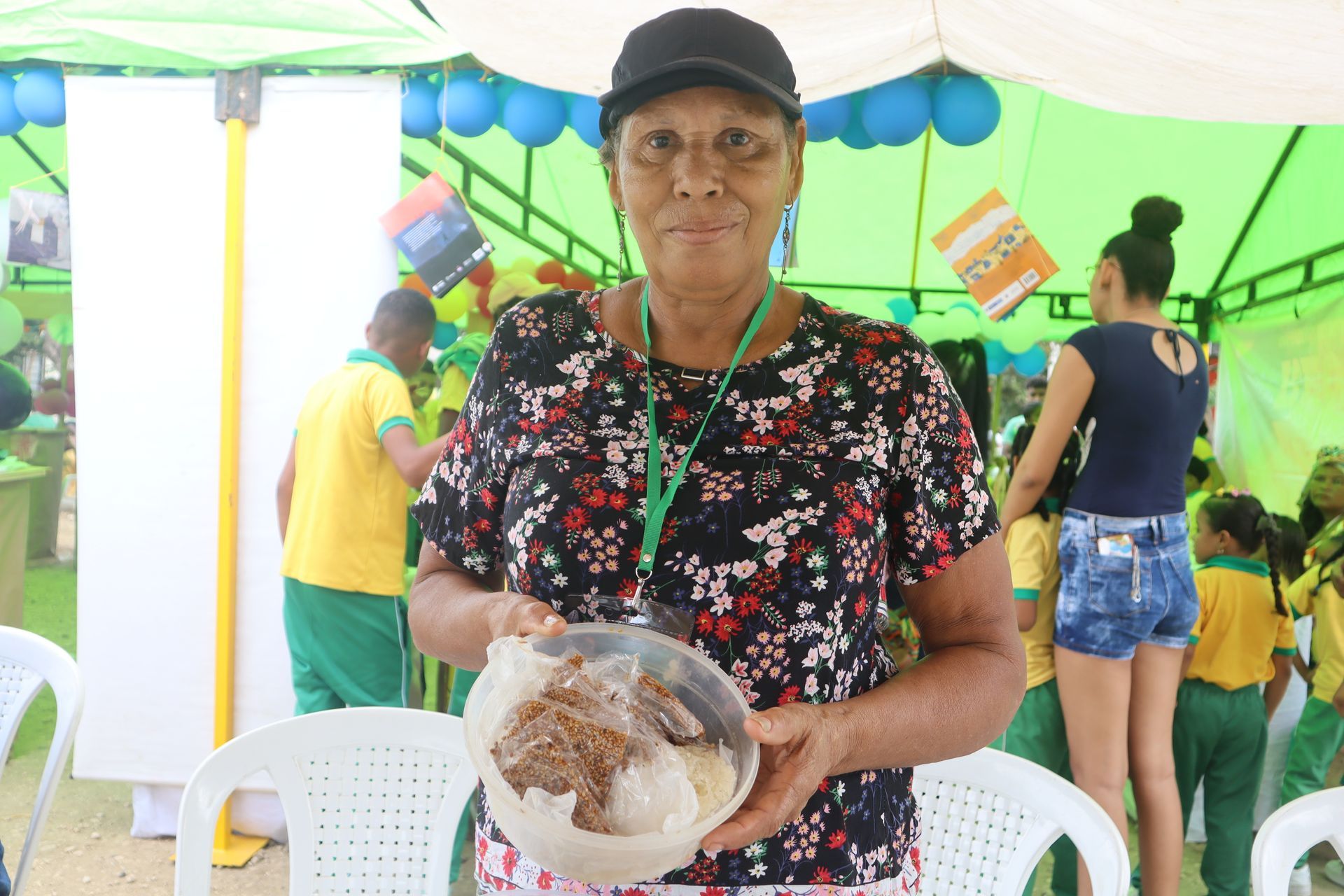 Mujer sosteniendo comida, posiblemente en un evento, sonriendo, vistiendo una gorra y una camisa floreada, al aire libre.