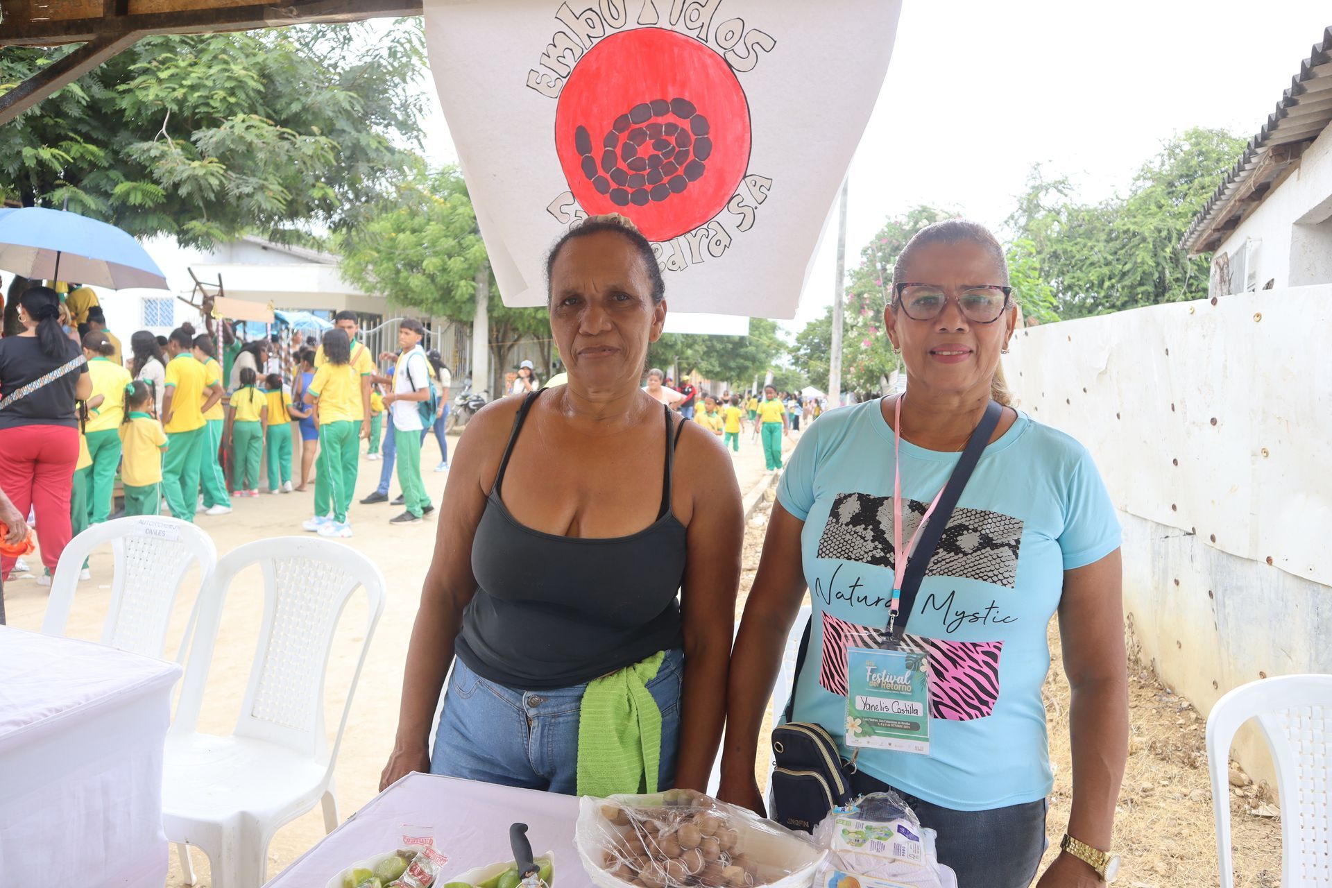 Dos mujeres están de pie ante una mesa, sonriendo, con una pancarta detrás de ellas y niños al fondo.