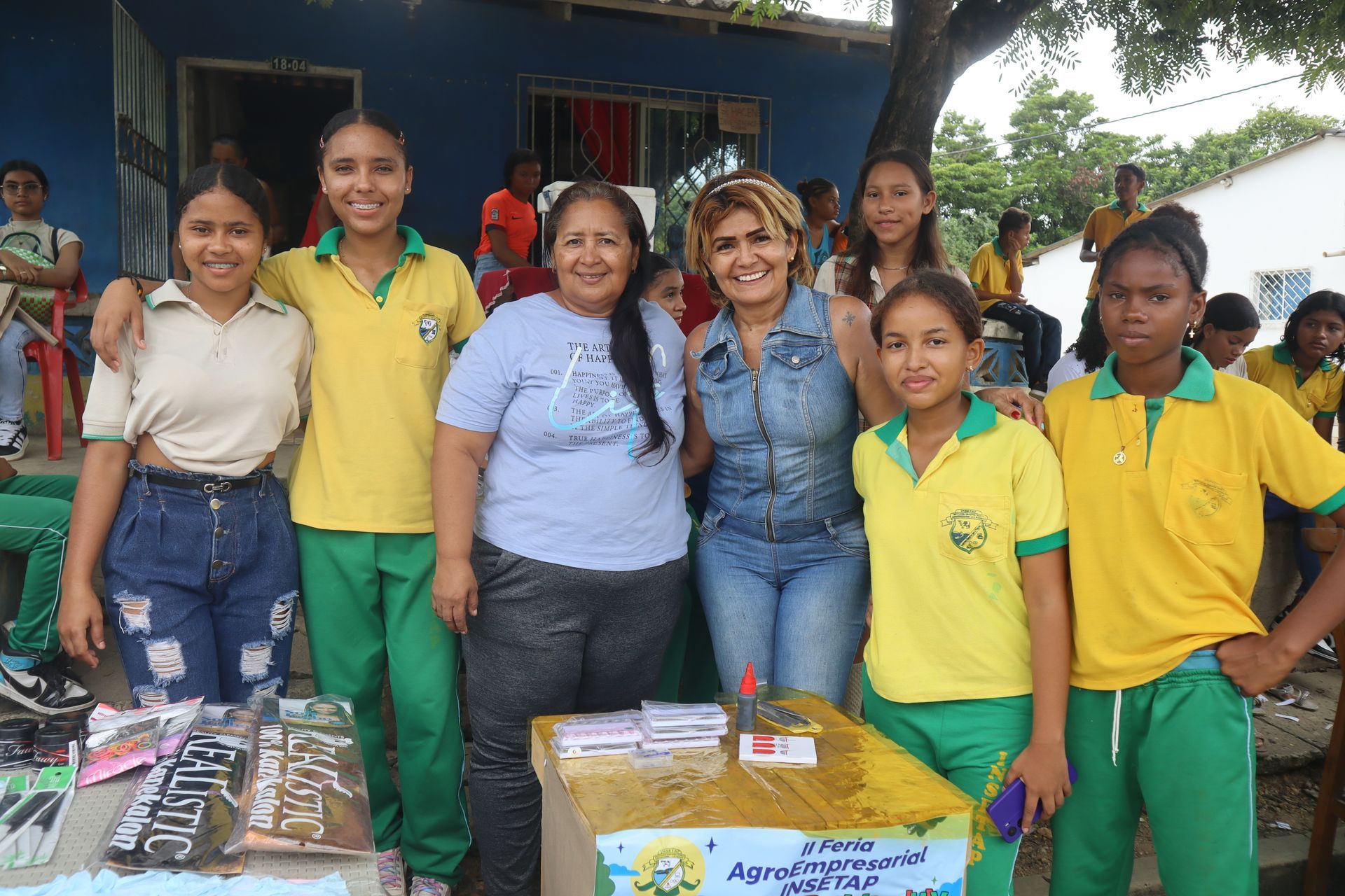 Grupo de mujeres y niñas sonriendo en un evento al aire libre con una mesa de útiles escolares.