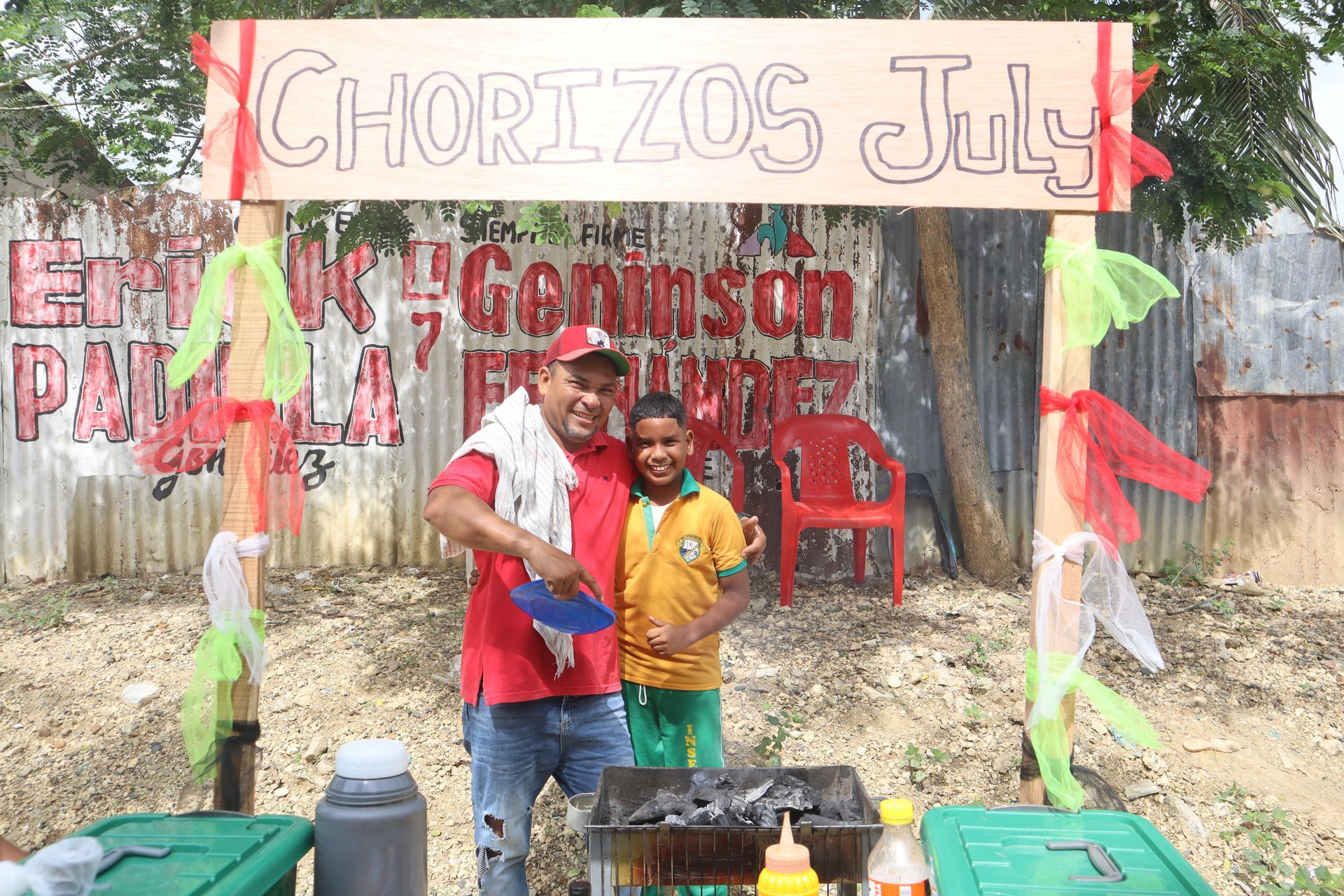 Un hombre y un niño junto a una parrilla de chorizos, bajo un cartel que dice 