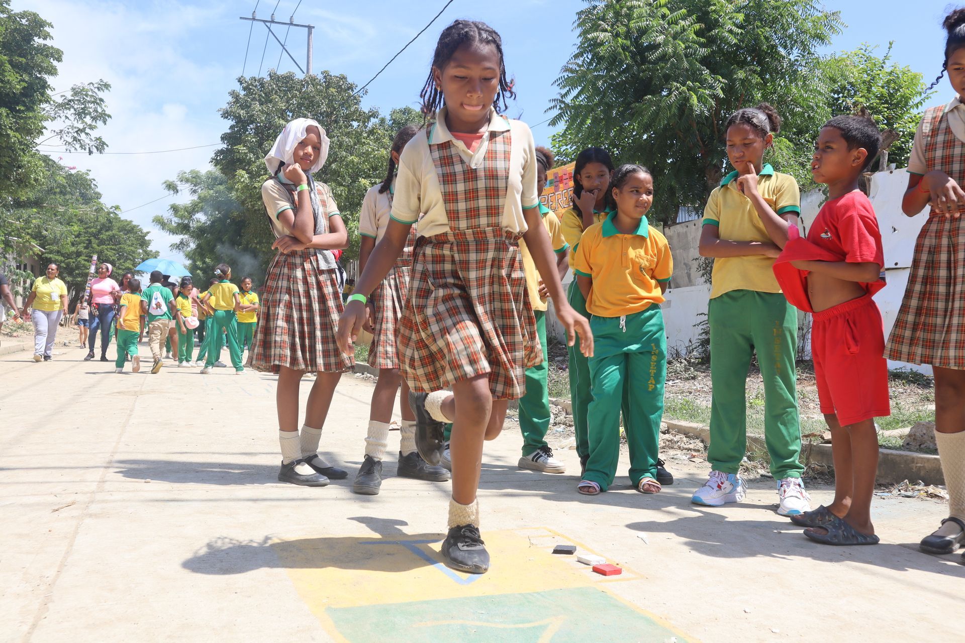 Niña jugando a la rayuela al aire libre, otros niños observan. Uniformes marrones, día soleado.