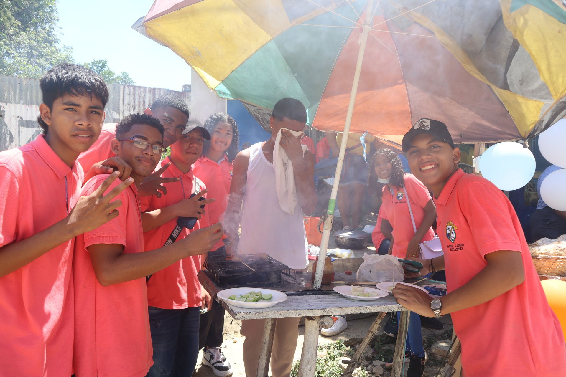 Grupo de adolescentes con camisetas rojas en un puesto de comida callejera bajo una sombrilla colorida; día soleado.