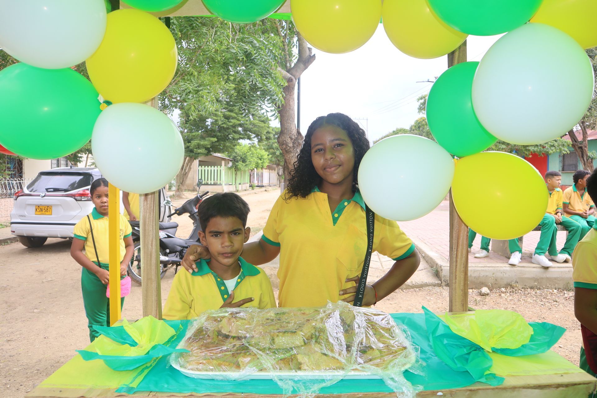 Dos personas detrás de un puesto de comida decorado con globos. Verdes, blancos y amarillos. Sonriendo, de pie frente a la comida expuesta.