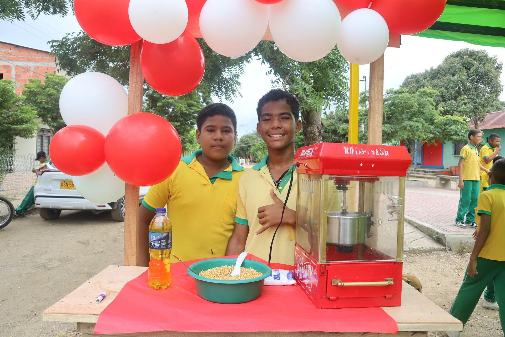 Dos niños sonriendo en un puesto de palomitas de maíz, con globos encima.
