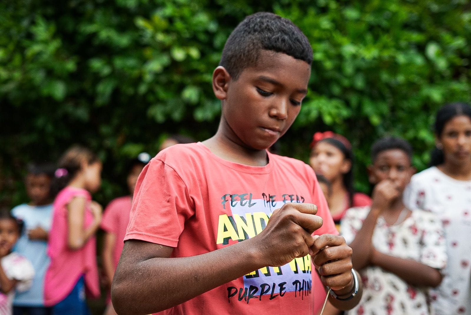 Niño con camisa roja mirando hacia abajo, posiblemente jugando con una cuerda, rodeado de niños.