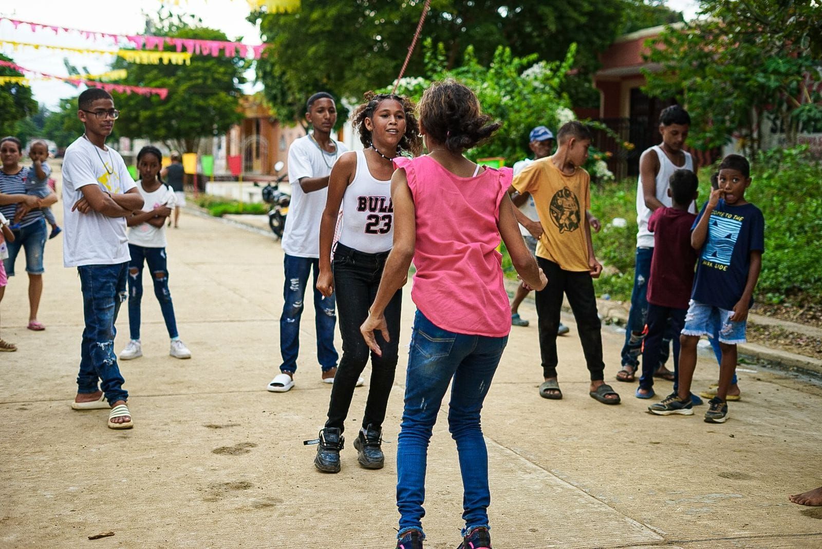 Niños jugando en la calle; una niña frente a otra, otros niños observando. Edificios y árboles al fondo.