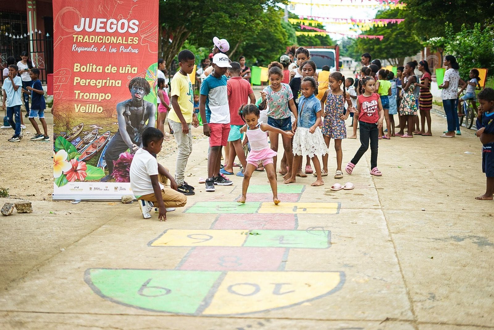 Niños jugando a la rayuela en una calle durante un evento; pancarta colorida en el fondo.