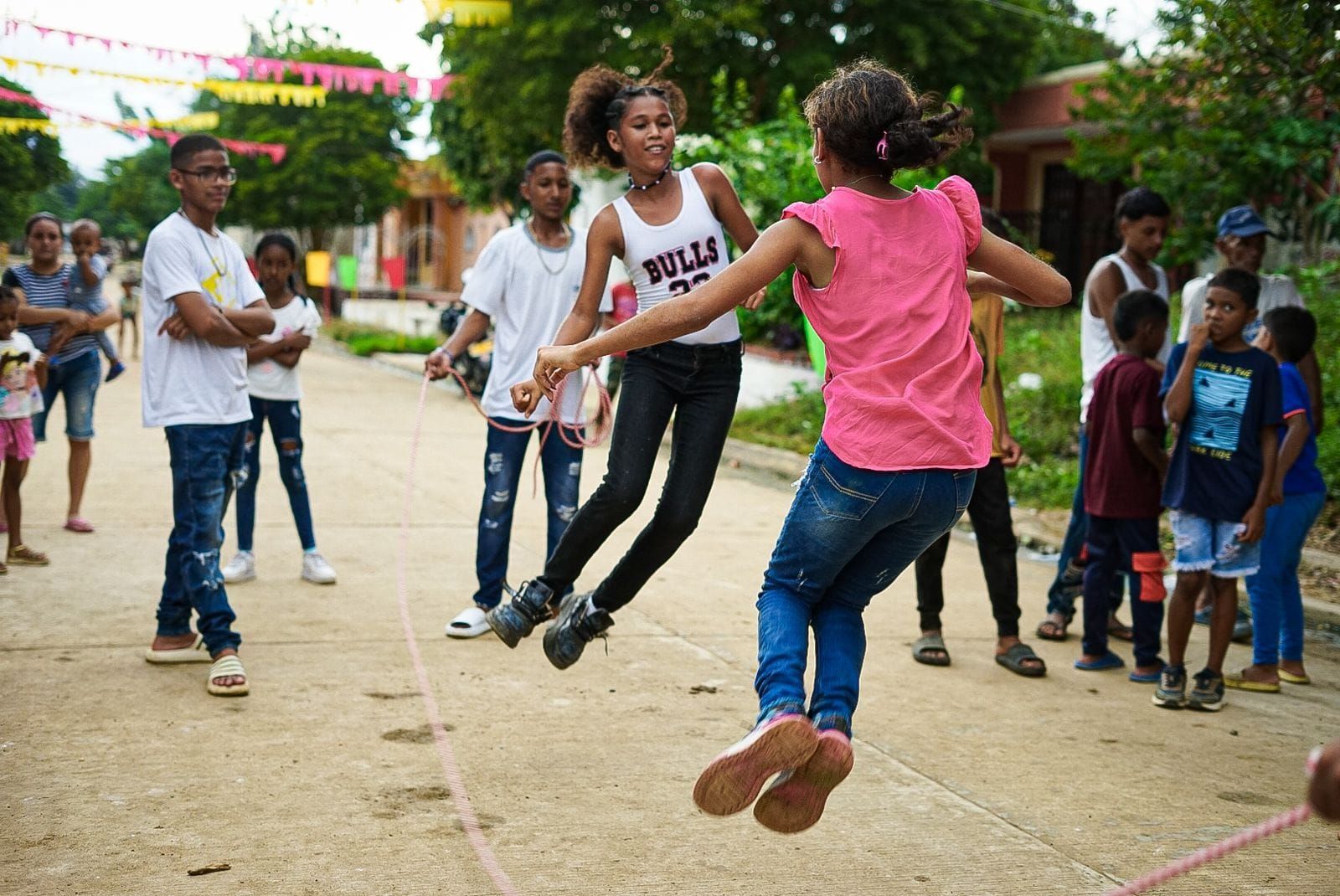 Niños jugando a saltar la cuerda en una calle; decoraciones festivas y espectadores al fondo.
