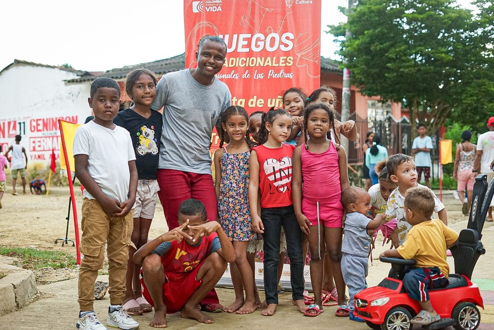 Un hombre posa con niños frente a un cartel de los Juegos Nacionales. Sonríen y están al aire libre.