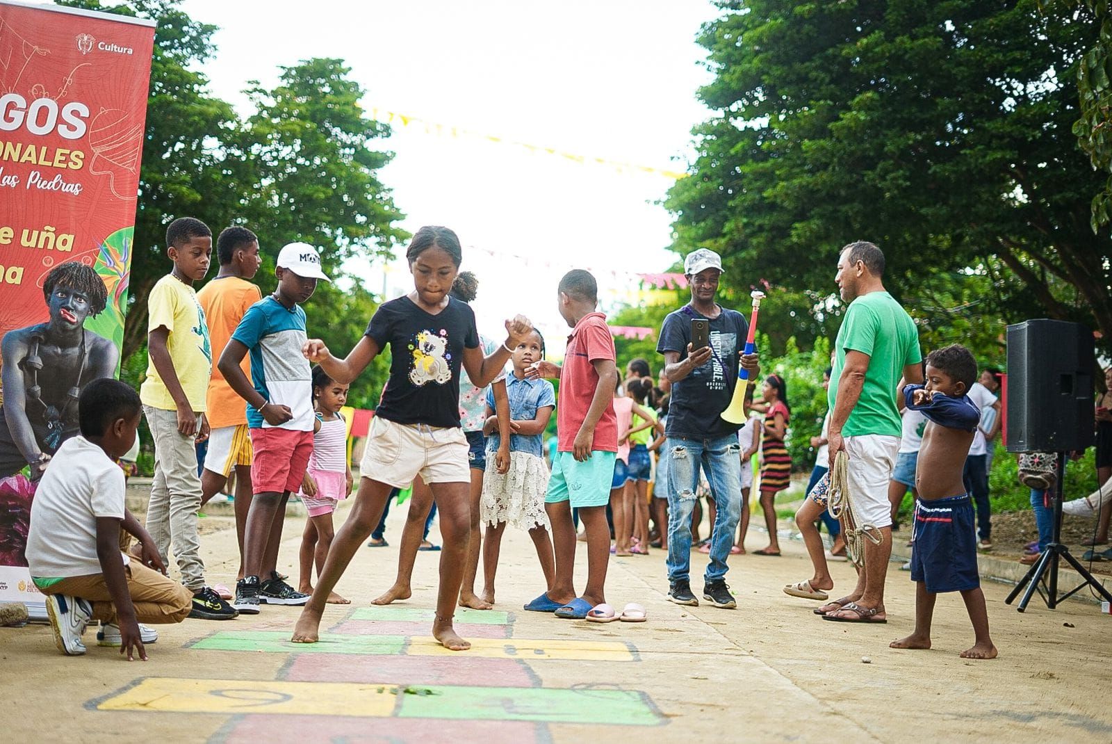 Niños jugando a la rayuela al aire libre, algunos observan. Personas de diversos tonos de piel, día soleado.