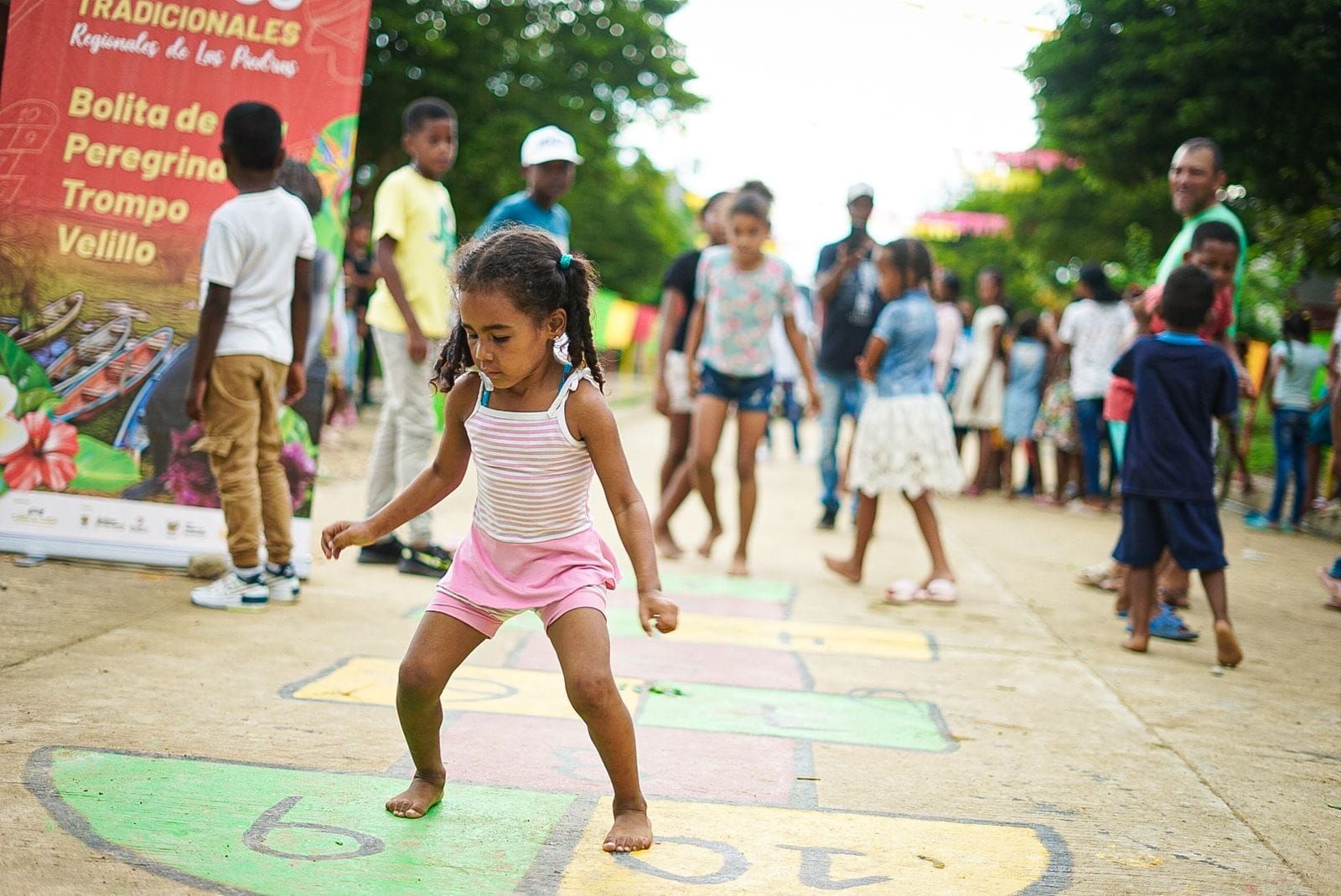 Niña jugando a la rayuela al aire libre, otros niños y adultos observando.