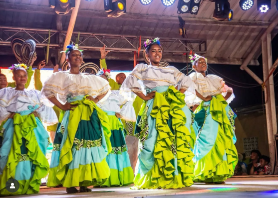 Bailarines con blusas blancas y faldas coloridas con volantes actúan en un escenario; de noche.