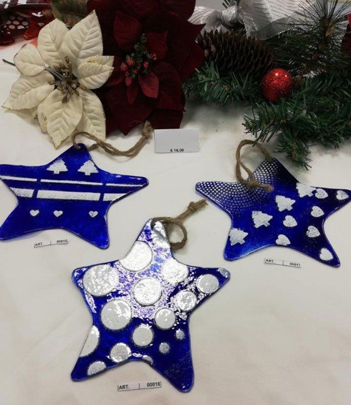 Decorazioni a forma di stella in vetro blu con disegni bianchi, esposte vicino a stelle di Natale e rami di pino.