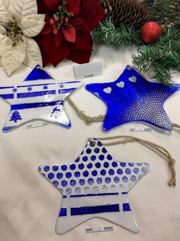 Tre decorazioni in vetro a forma di stella blu e bianca con motivi diversi, appese a un filo naturale. Decorazioni natalizie sullo sfondo.