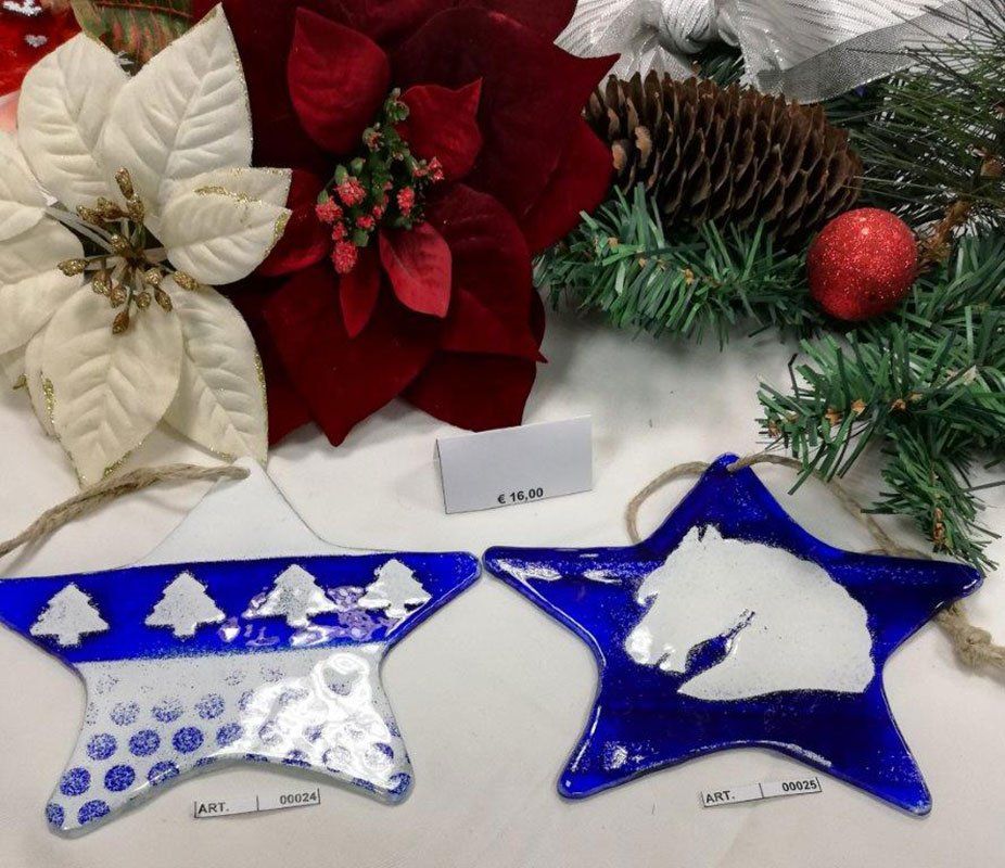 Due addobbi natalizi a forma di stella blu con disegni bianchi, circondati da stelle di Natale e pini, e decorazioni.