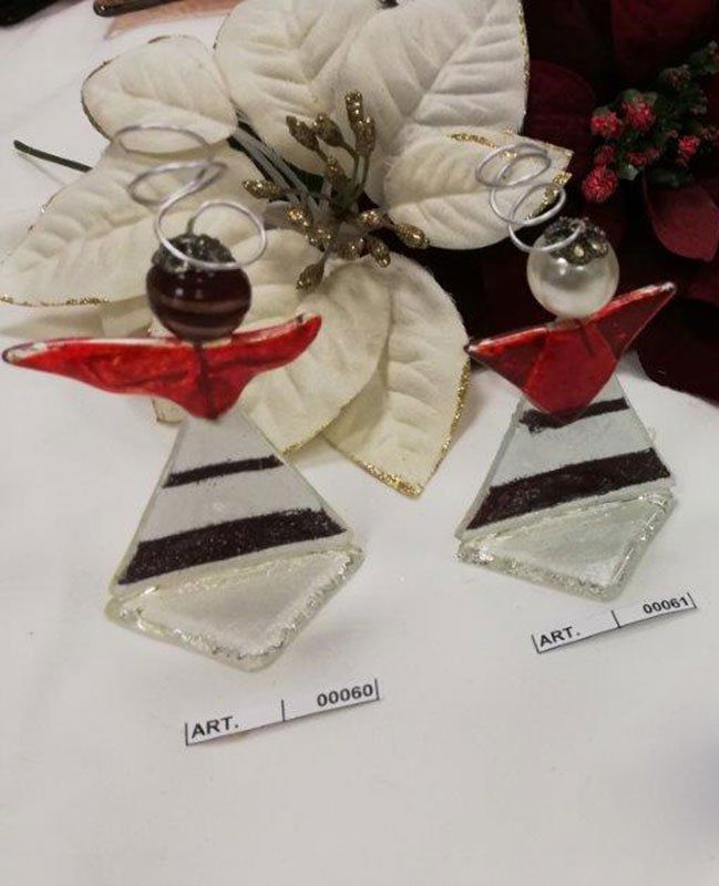 Due ornamenti in vetro a forma di angelo con dettagli bianchi, rossi e neri, vicino a fiori di stelle di Natale.