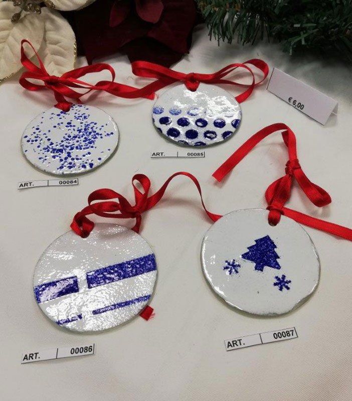 Quattro decorazioni natalizie in ceramica bianca con disegni blu e nastri rossi, esposte su una superficie bianca.