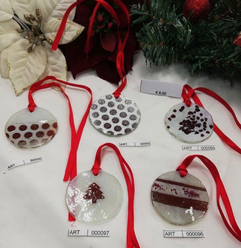 Primo piano di cinque decorazioni natalizie rotonde in vetro con nastro rosso, vari motivi su sfondo bianco.