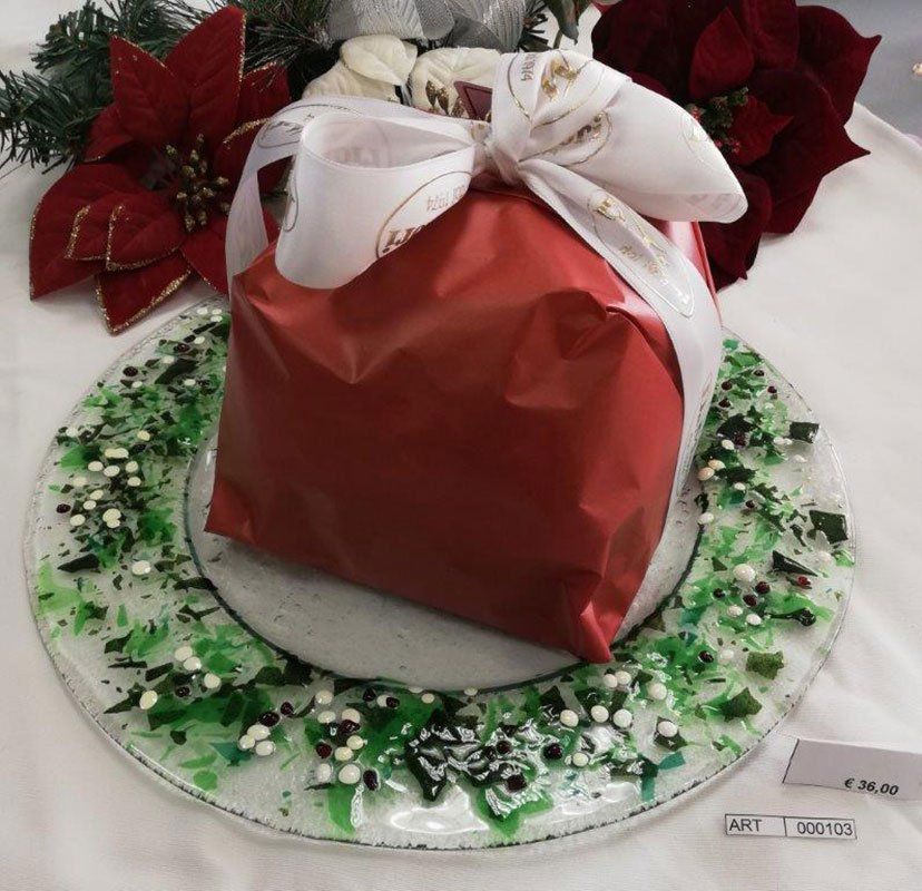 Il regalo, confezionato in rosso e legato con un fiocco bianco, è appoggiato su un piatto di vetro decorativo con decorazioni verdi, bianche e nere.