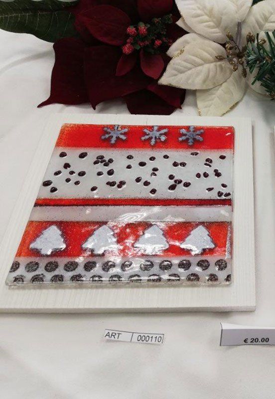 Opera d'arte in vetro fuso con motivi natalizi rossi, bianchi e neri, su un supporto bianco, con sfondo floreale.