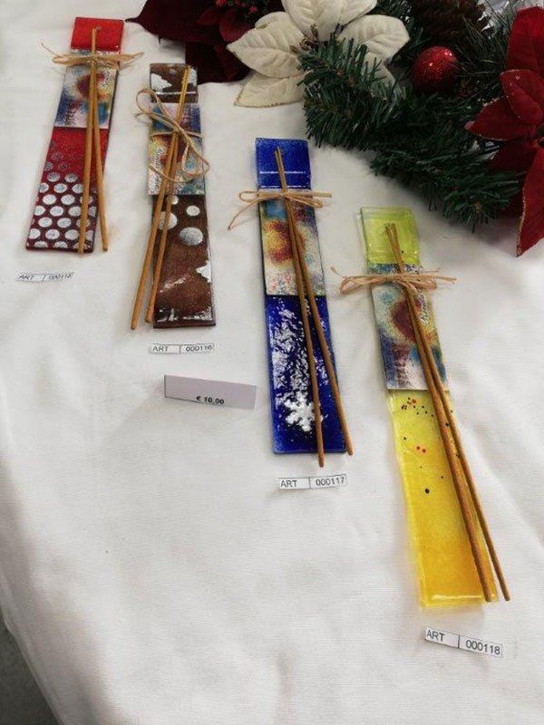 Quattro porta incenso colorati in vetro su una superficie bianca, decorati con spago e bastoncini di incenso.