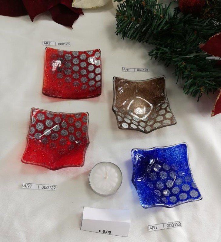 Quattro portacandele quadrati in vetro colorato con motivi a pois argentati, accanto a una candela e decorazioni natalizie.