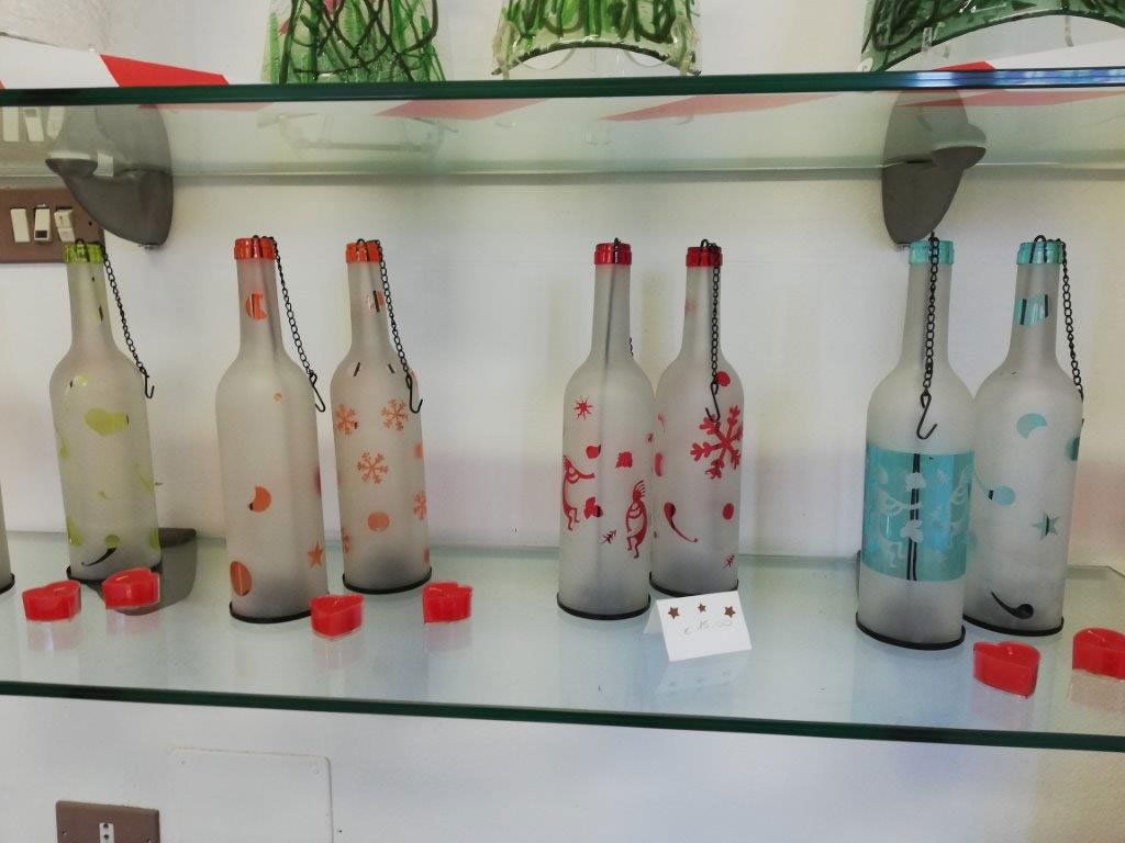 Mensola espositiva in vetro con bottiglie smerigliate decorate con motivi rossi e arancioni e candele votive rosse.