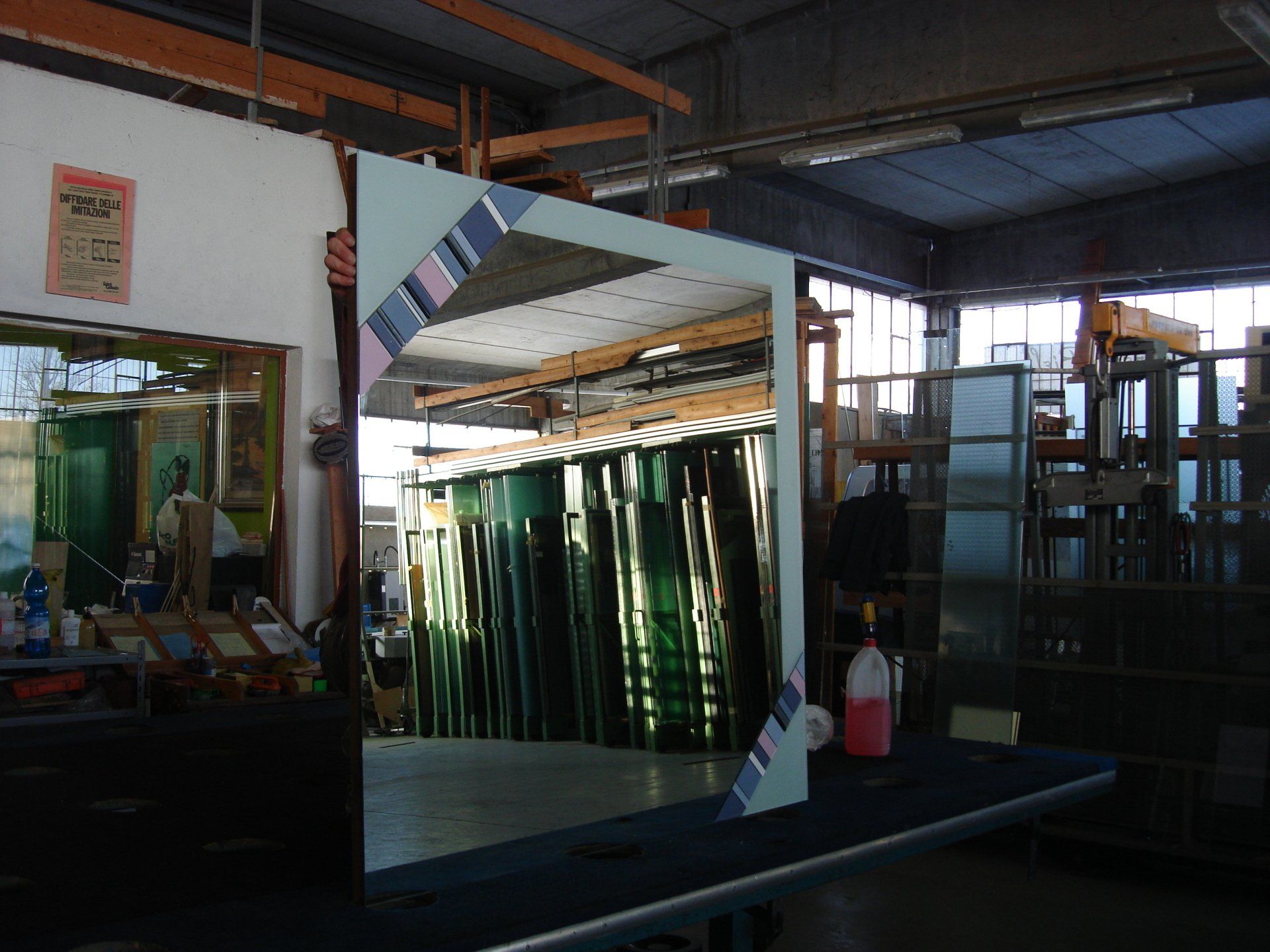 Un grande specchio è appoggiato su un tavolo in un laboratorio. Sullo sfondo sono impilate lastre di vetro.